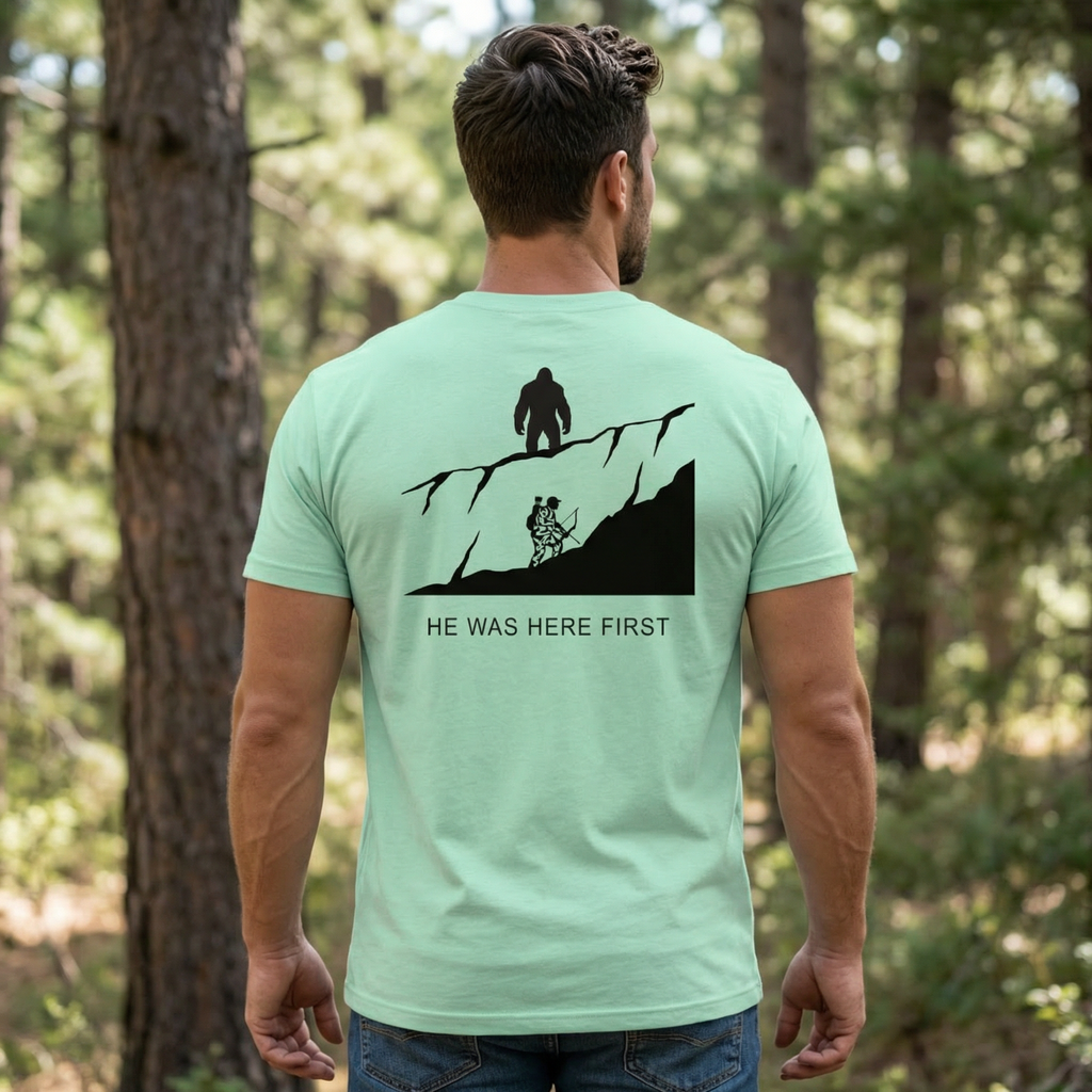 Back of man wearing He Was Here First Bigfoot Hunting Tee in Heather Mint featuring a ridge line Bigfoot graphic above a climbing bowhunter in a forest setting.
