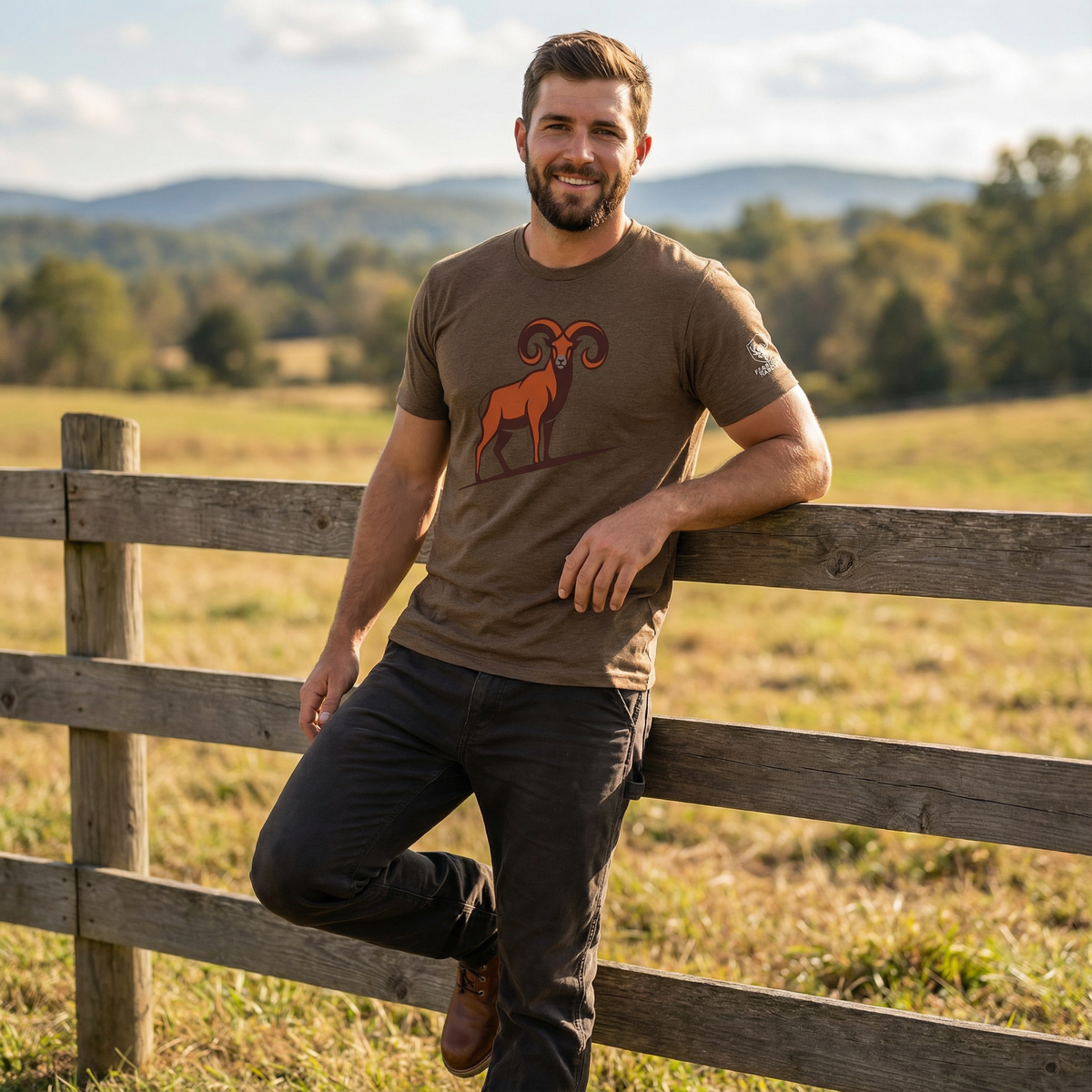 Man leaning on a wooden fence in a scenic outdoor setting. Front left lifestyle view of a man wearing the heather brown Full Curl Ridge Eternal Core Tee, showing the tee in motion with a relaxed fit against a high country, mountain-inspired backdrop.