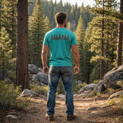 Lifestyle back image of the Men’s Fade Into the Pines Bigfoot Core Tee in Teal highlighting the clean back graphic of a walking Bigfoot silhouette moving through a pine tree line beneath the words fade into the pines.