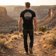 Model wearing the Mens Hunt the Ages Premium Everyday Tee in solid black showing the back prehistoric triceratops and elk graphic in a natural outdoor setting.