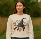 Woman wearing a beige sweatshirt with a graphic design of a witch hat, skull, and broomstick in a natural setting.