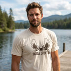 Man outdoors wearing the Men's Paddle Country Moose Tee in Heather Dust front view showing the paddle antler timber graphic in a lake setting.